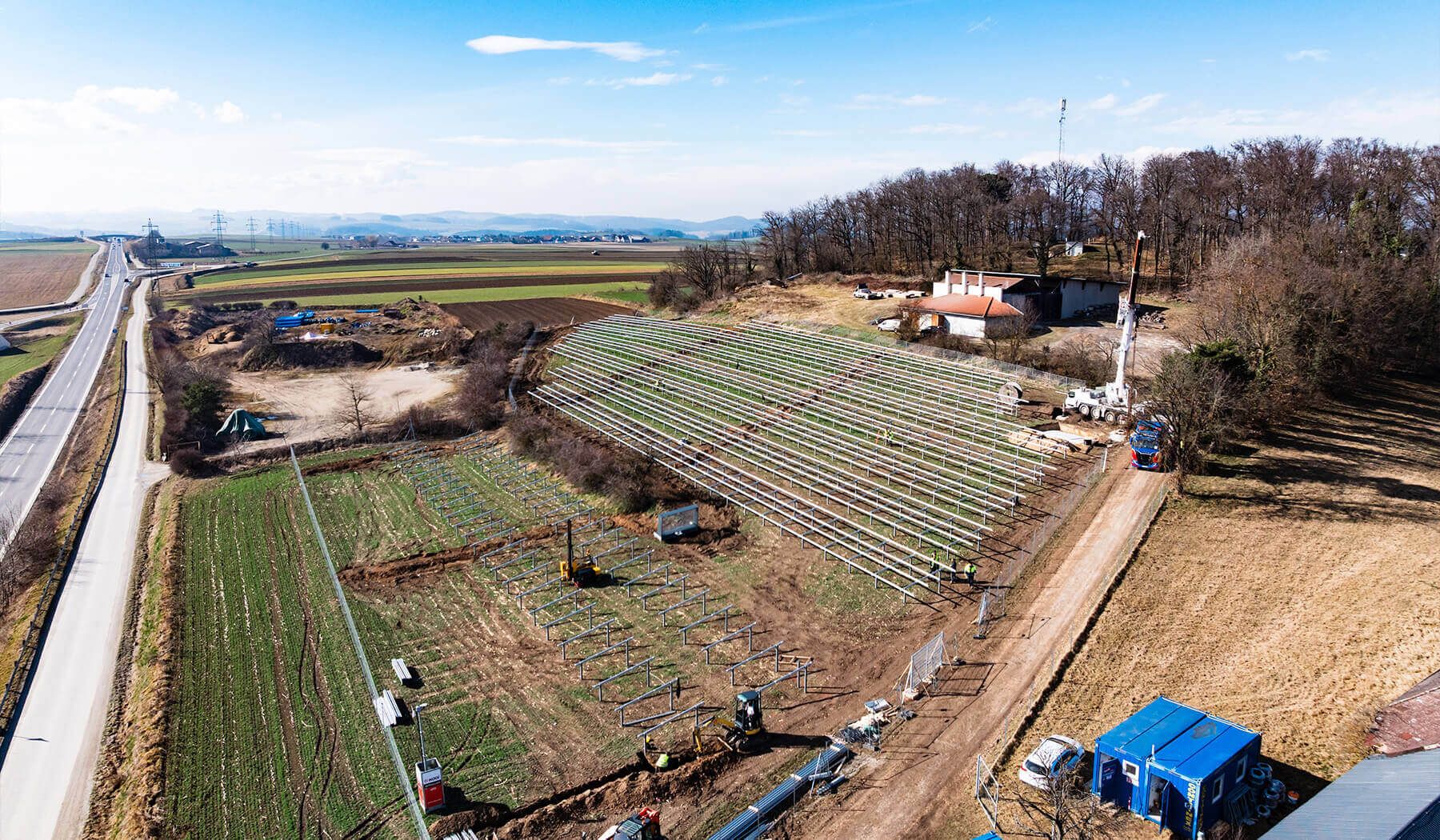 Luftaufnahme einer Photovoltaik-Freiflächenanlage im Bau, mit aufgestellten Unterkonstruktionsreihen neben einer Straße und landwirtschaftlichen Flächen.