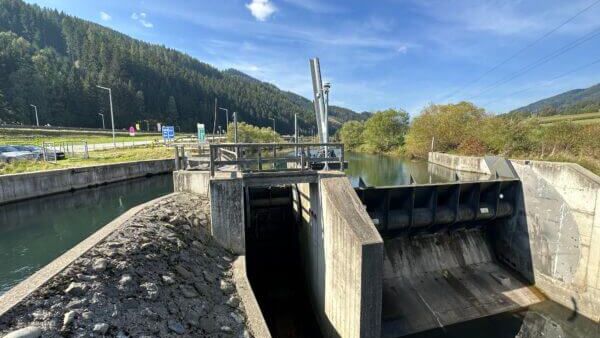 Damm mit Schleusentor an einem Fluss, umgeben von bewaldeten Hügeln.